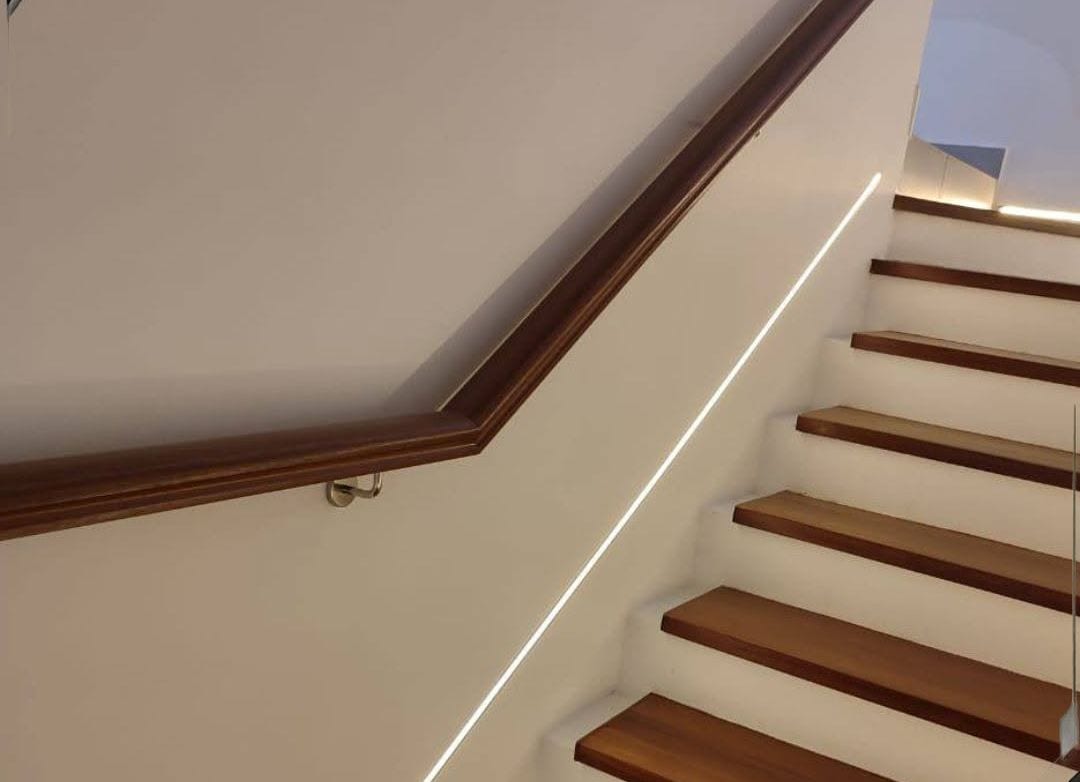 A dark timber wall handrail seen from the top landing, with a lit line along the stringer wall and timber treads below.